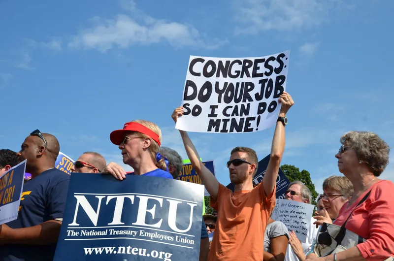 A group of people gather outside holding signs. One sign says “Congress: Do your job so I can do mine!” Another sign says “N T E U The National Treasury Employees Union. www.nteu.org”