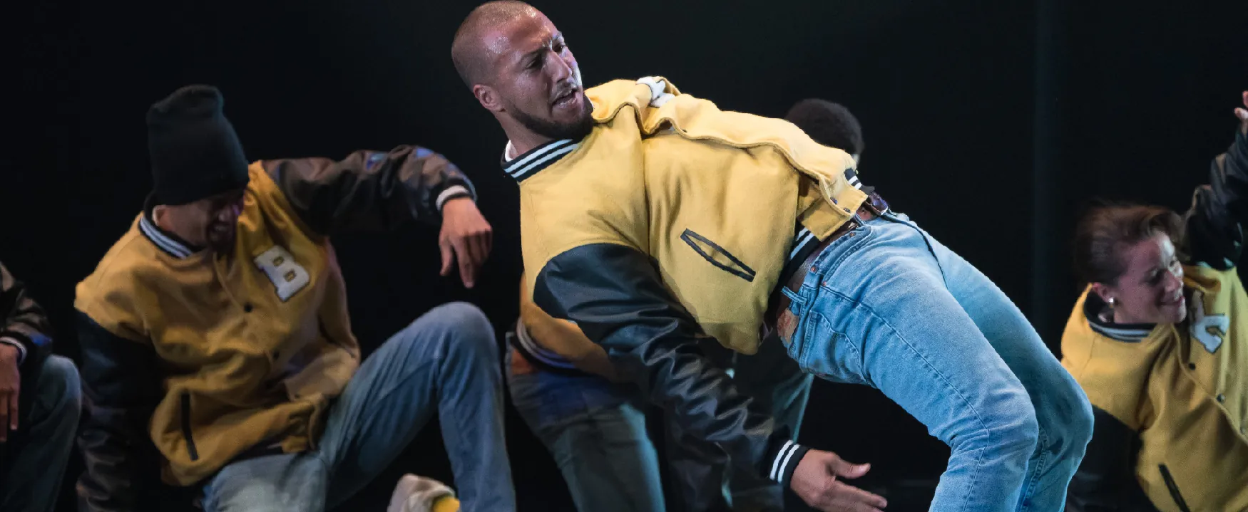 A photo shows a group of dancers rehearsing on a stage. In the foreground, a man can be seen leaning far backward with his back positioned almost parallel to the floor. Others in the background tend to fall while attempting to do the step.