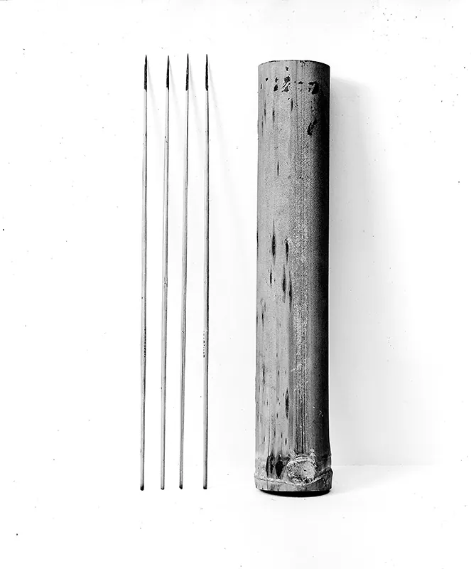 A cylinder-shaped storage container is next to four long narrow darts with darkened tips.