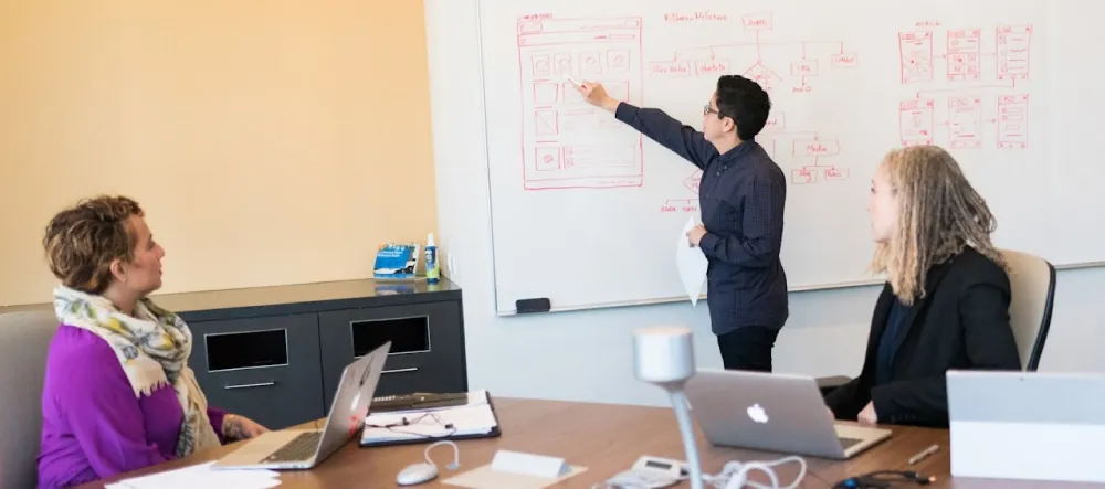 Photo of individual pointing to drawings on whiteboard, while two other individuals look on while seated with open laptops on table.