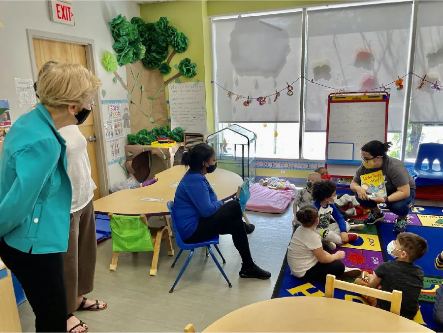 Photo of childcare center staff reading a book to children sitting on a rug, with other adults looking on in classroom type setting.