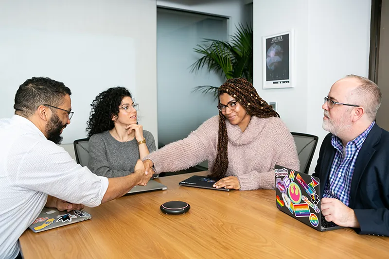 Four business people sit around a conference table with laptop computers in front of them. Two people are reaching across the table, shaking hands.