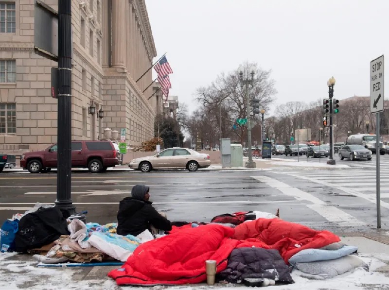 An individual sits on a street corner; the road is wet and there is a dusting of snow on the sidewalk. A sleeping bag and other personal effects surround the individual on the sidewalk.