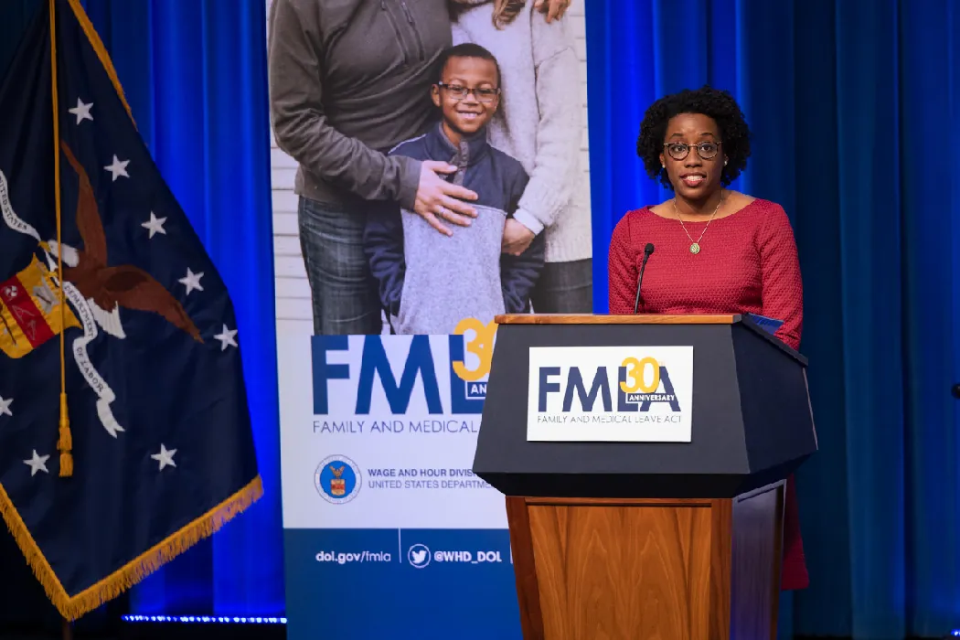 U S Representative Lauren Underwood stands behind a podium that says FMLA. At left is a larger banner with the words FMLA and a picture of a child with two adults as well as a flag with the seal of the United States.