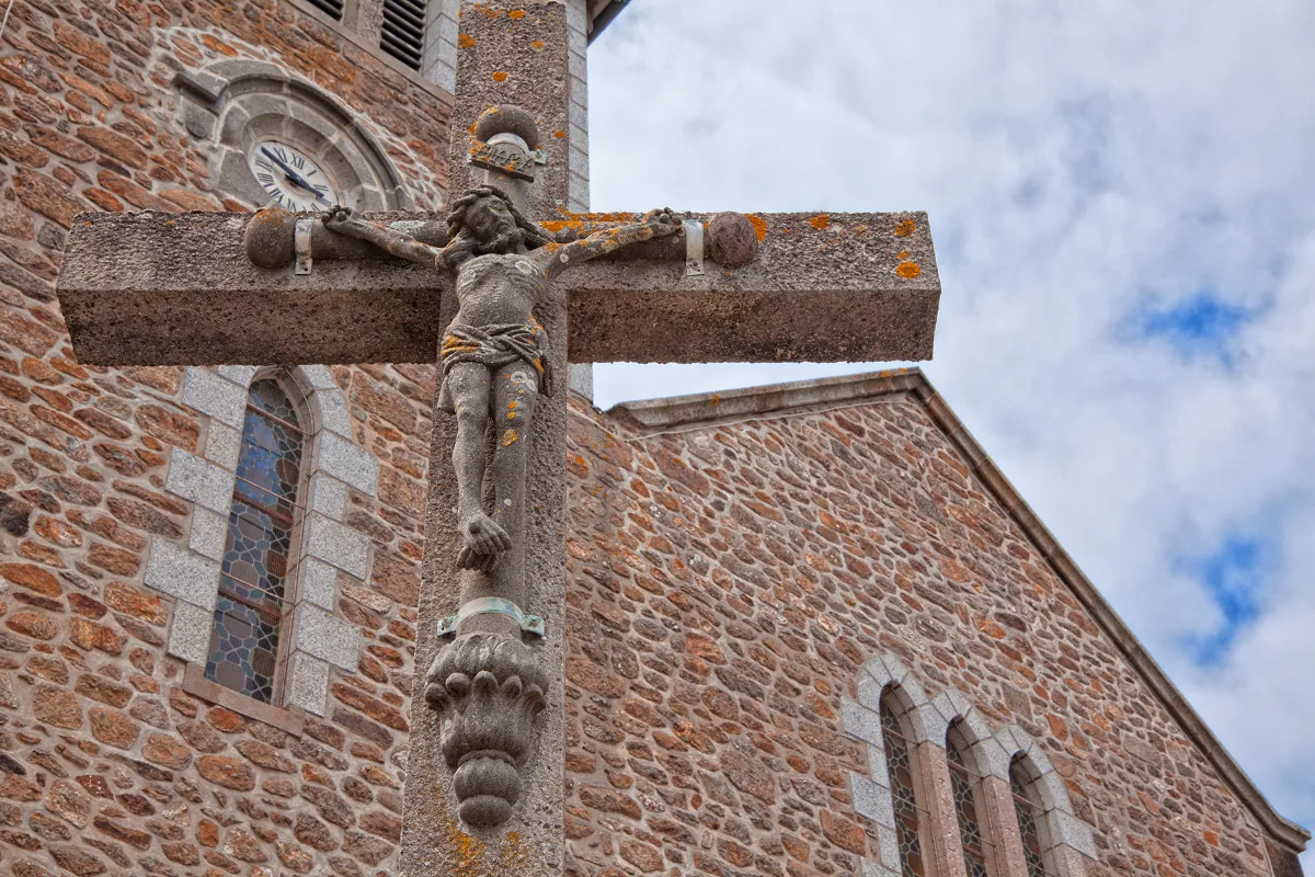 Photo of a stone cross with Jesus crucified on it with a brick church in background.
