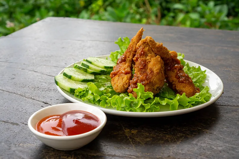 A plate of fried chicken wings sits on a bed of lettuce with a few cucumber slices on the edge of the plate. A small ramekin containing ketchup and chili sauce is next to the plate. Both the plate and the ramekin are on a wooden table.