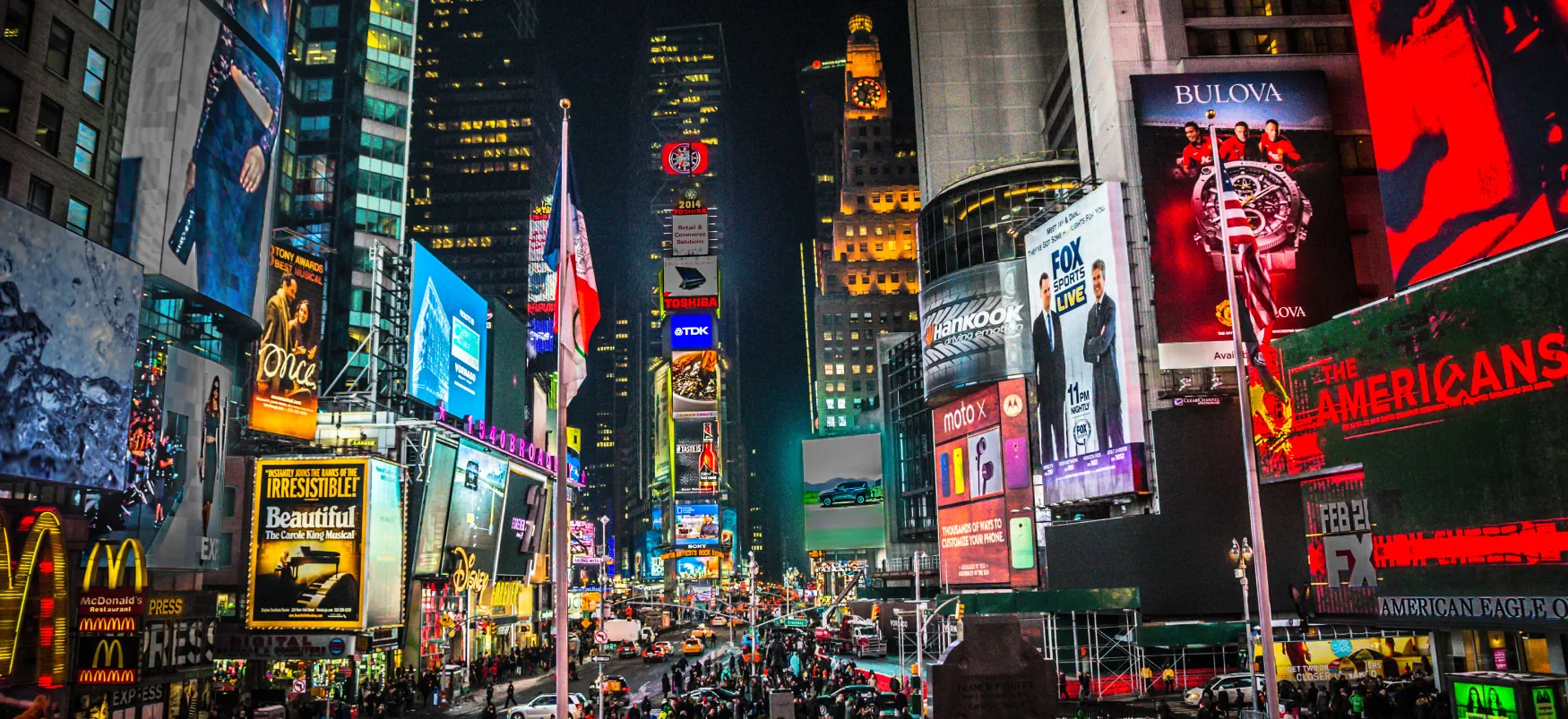 Times Square in New York City is shown at night. The tall skyscrapers are lit up. Electronic billboards show ads for T V shows, Broadway plays, products, and restaurants. The streets are crowded with people and traffic.