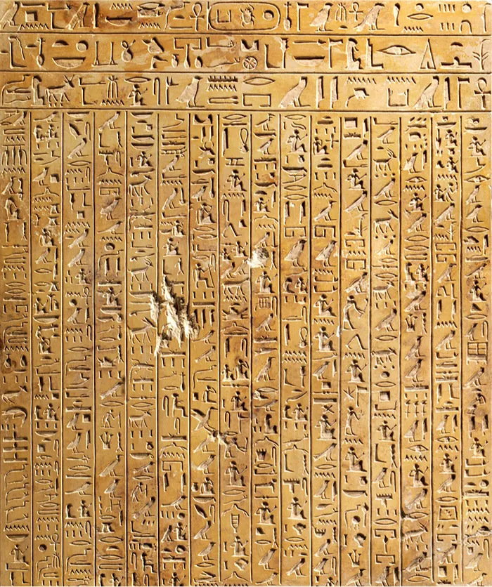 A picture of a brown rectangular stone is shown with a red and beige checkerboard trim around the top and two sides of the stone. Three rows run across the top and sixteen columns run under the rows. All the rows and columns are filled with various hieroglyphics. Along the bottom the head of two men in helmets can be seen as well as a scarf with tassels in between them. Scratches and cracks can be seen throughout the stone.