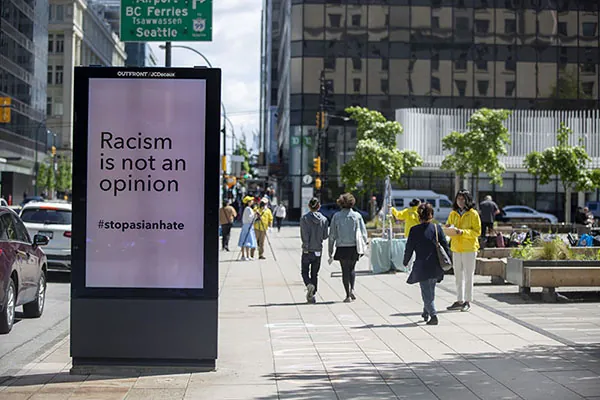 A sidewalk billboard reads “Racism is not an opinion. #stopasianhate.” A few people walk on the sidewalk.