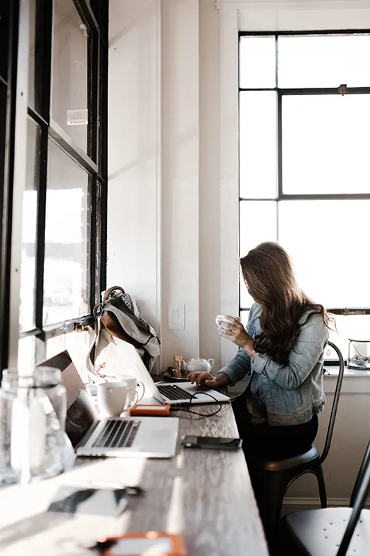 A person holding a cup sits at a table with several laptops and works at one of them.