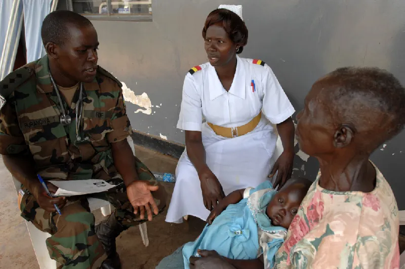 A person in a nurse's uniform sits next to a speaking person who wears camouflage and a stethoscope around their neck and holds papers and a pen. A third adult across from them holds a sleeping infant.