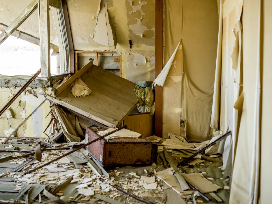 The interior of a room shows signs of destruction and decay, with peeling paint and wallpaper, broken furniture, and debris covering the floor.