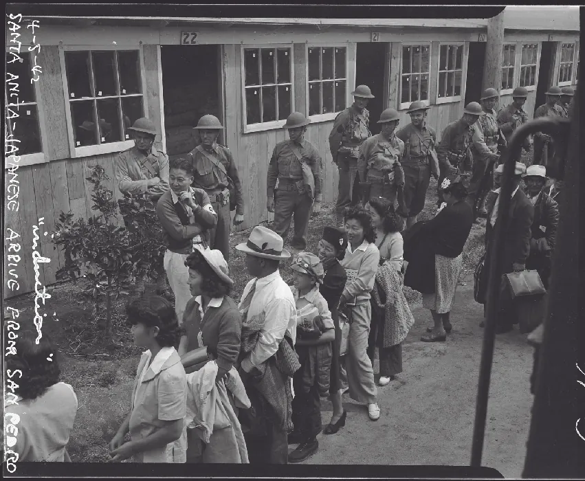 A group of Japanese American men and women stand in a line outside of a building. A row of soldiers in uniform stand off to the side watching them.