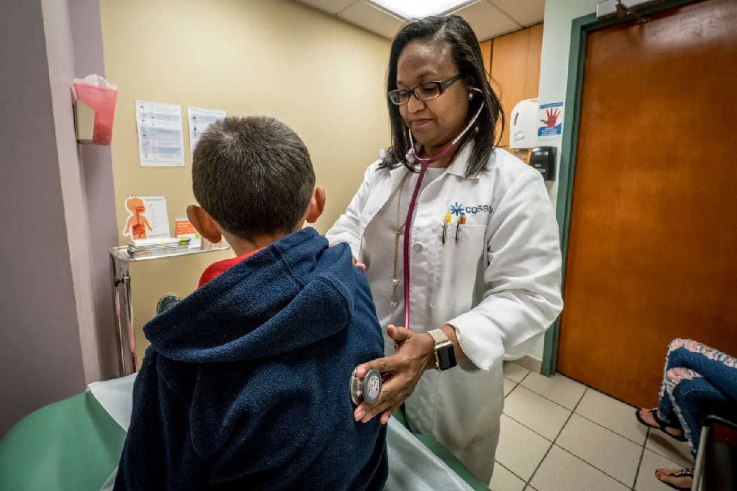 A child sits on an exam table in a doctor's office while a health care provider listens to the child's lungs with a stethoscope.