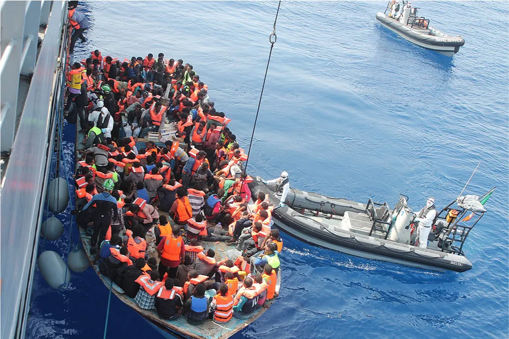 A picture is shown of boats in very calm, blue water. At the left is a metal rail above a tall silver ship hull. Three people wearing orange life jackets can be seen climbing a rope ladder from a flat, plain boat next to the ship. Two people wearing black life jackets over white hazmat suits are at the bottom of the rope ladder. Four gray buoys are wedged between the ship and the boat, tied to the large ship. The plain, flat boat is completely filled with people. The people inside are dark-skinned with dark hair, all wear various orange life jackets and sit crowded together. A person in a green helmet and blue/black wet suit stands on the edge of the crowded boat holding on to the buoy rope. To the right of the boat there is a white and gray Rigid Inflatable Boat (RIB) - a boat with a rigid hull and inflatable sides - with two people in white hazmat suits and black life jackets, one at the front of the boat and one driving in the back. Another RIB boat with the words “Naval Service” at the front can be seen in the distance with two hazmat-clad people toward the back.