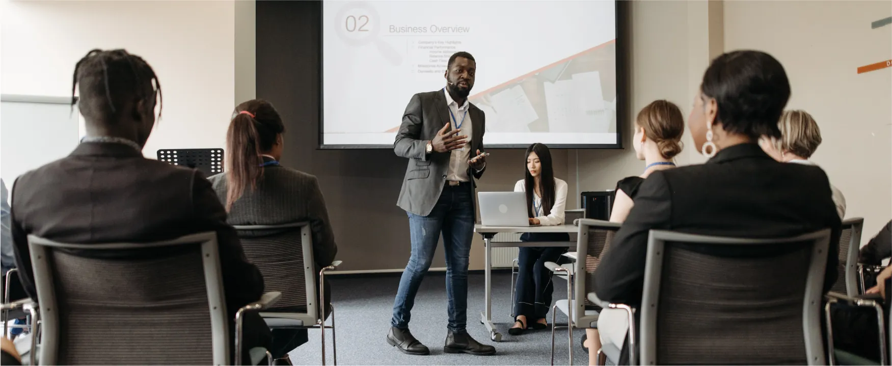 A man stands in front of a small audience while giving a presentation. There is a slideshow being projected behind him.