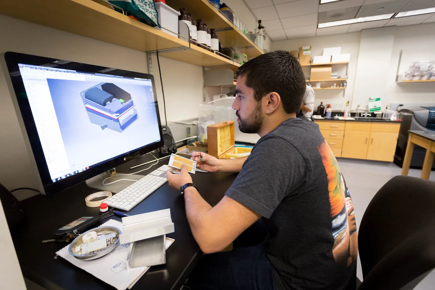 A person in a workspace holds a small component while looking at a screen with a detailed 3D rendering of a more complex design. 