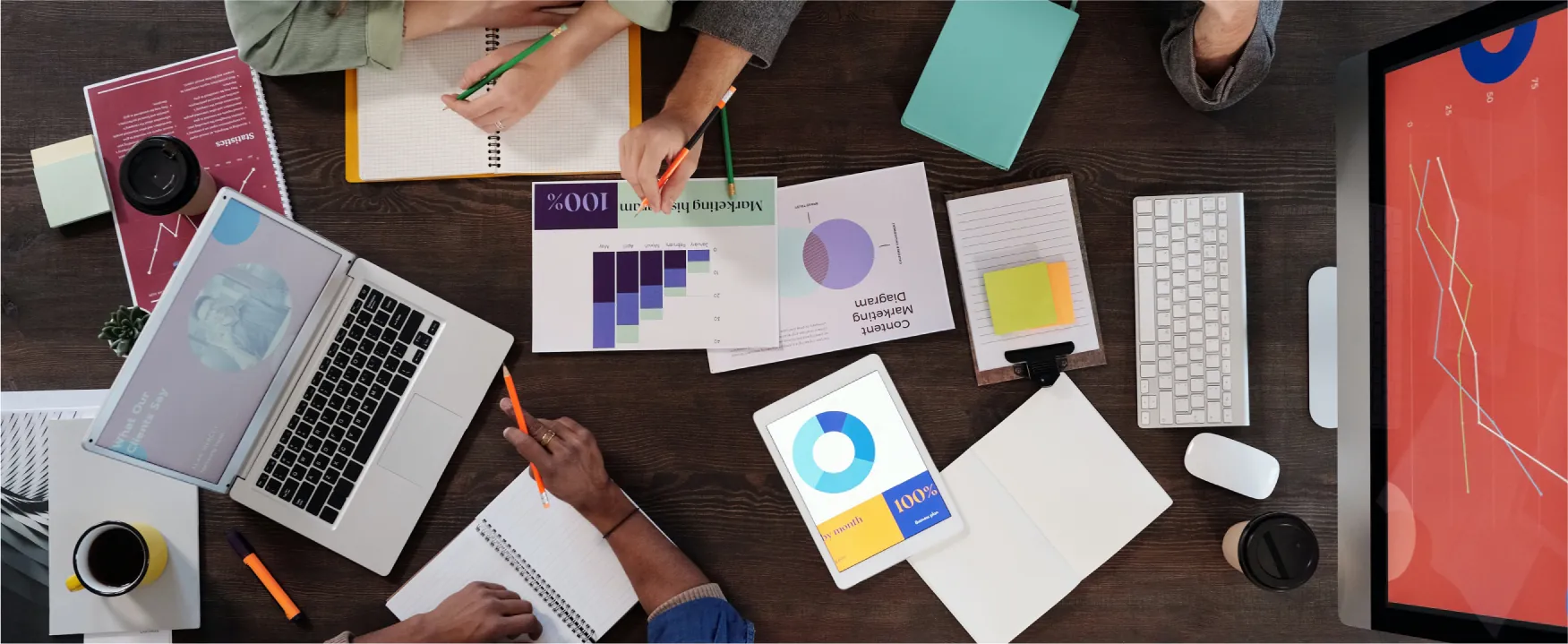 A desk with papers, computers, tablets, coffee cups, pencils, markers and two people’s hands is visible from above.