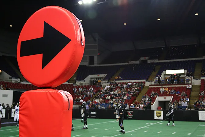 A large orange circle with an arrow printed on it facing to the right atop a rectangular base, known as chains in football, is shown prominently in the foreground.  In the background there is a game of football ongoing with fans in their seats.