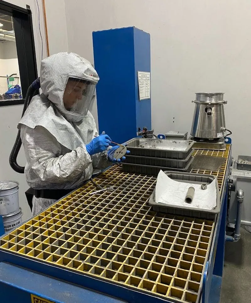 A person wearing a robust protective suit with air hoses emerging from it is holding a small part and a device. The workstation consists of a table with a grid on top rather than a solid surface. Multiple trays holding parts and other objects are nearby. 
