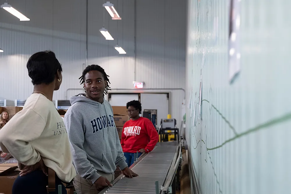Several people stand along a roller conveyer in a large warehouse-like space with boxes nearby.