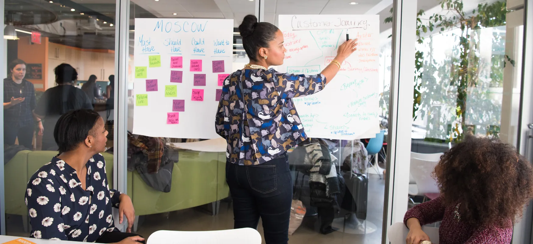 Photo of individual pointing to notes on white paper taped to window, while two other seated individuals look on in an office setting.