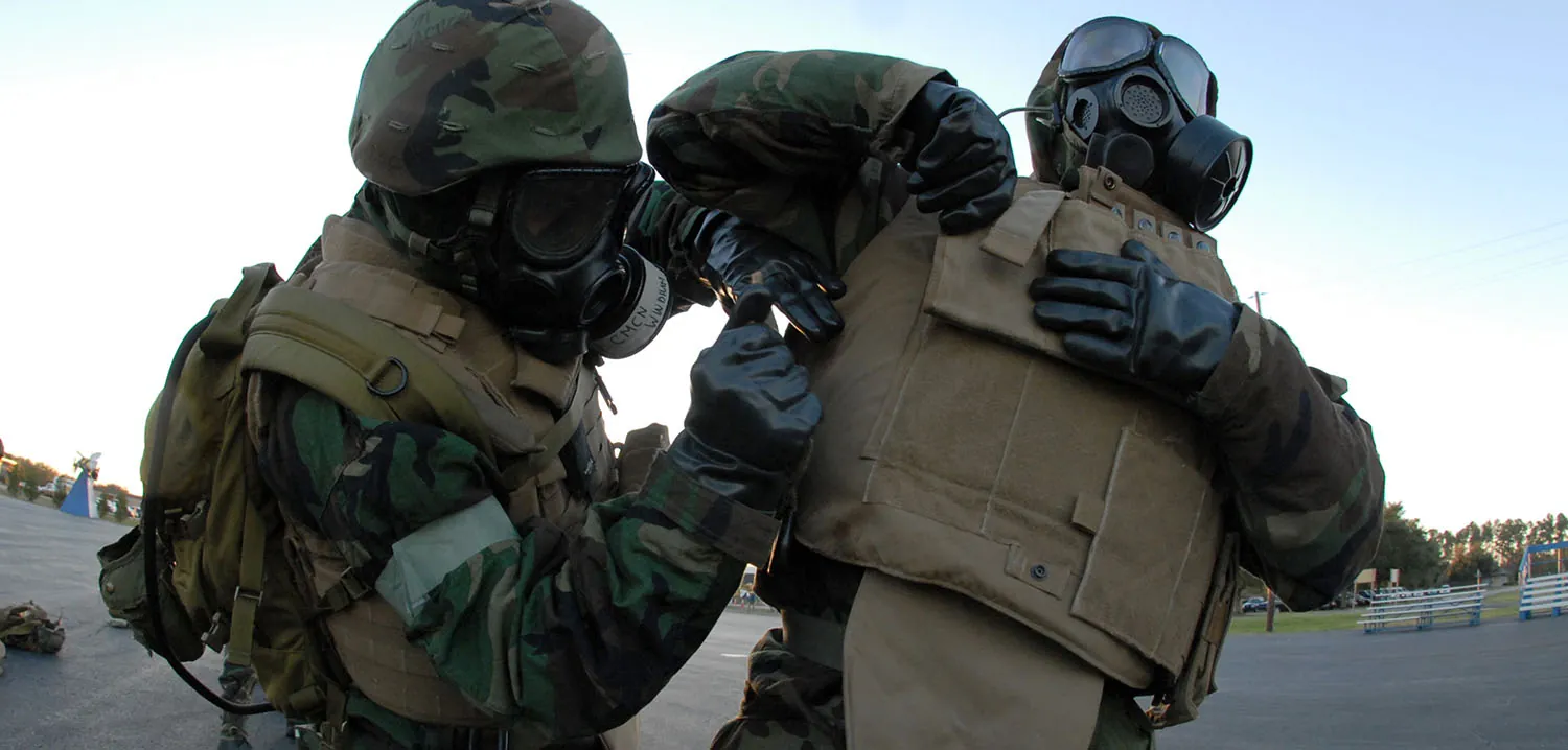 A soldier adjusting the straps on the bulletproof vest of another soldier. Both soldiers are in uniform, wearing helmets, gas masks, and gloves.