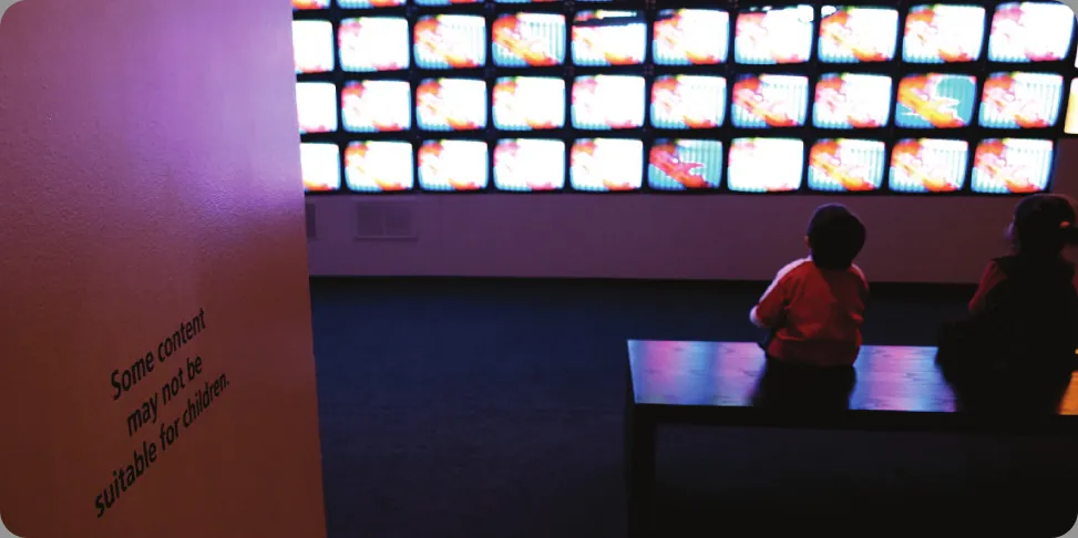 Children sit in front of a bank of television screens. A sign on the wall says, “Some content may not be suitable for children.”