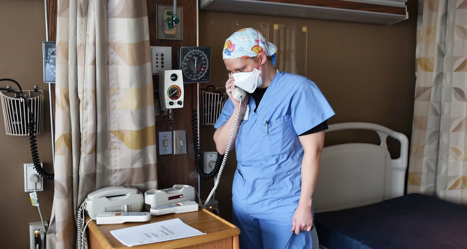 A color photograph shows a nurse standing in a hospital room holding a phone to her ear.