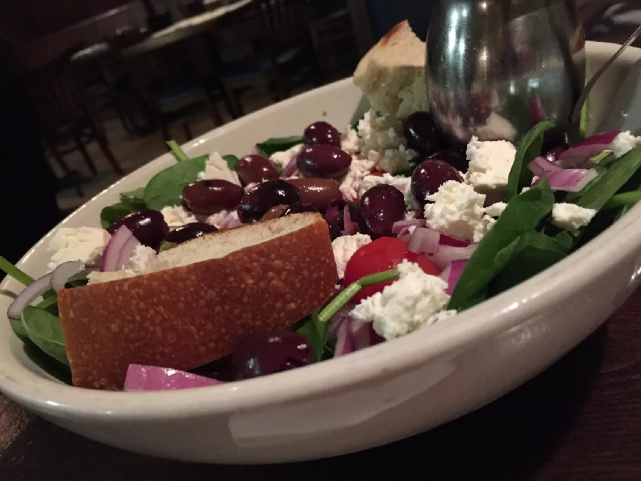 A small bowl contains leafy greens, red onion slices, olives, tomatoes, and crumbled feta cheese. There is a slice of bread in the bowl and a small silver dressing carafe.