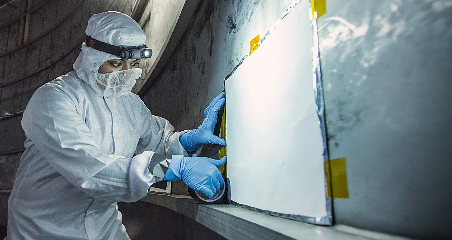 A person wearing a coverall and face mask attaches a rectangular panel to a wall.