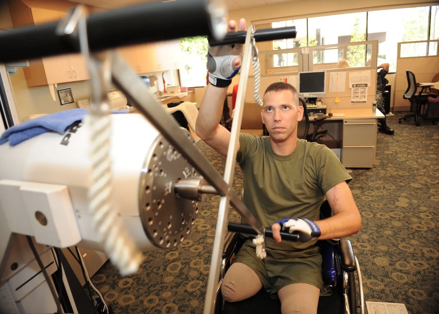 Man in a wheelchair wearing workout gloves using an exercise machine in an office setting, focusing on rehabilitation exercises.
