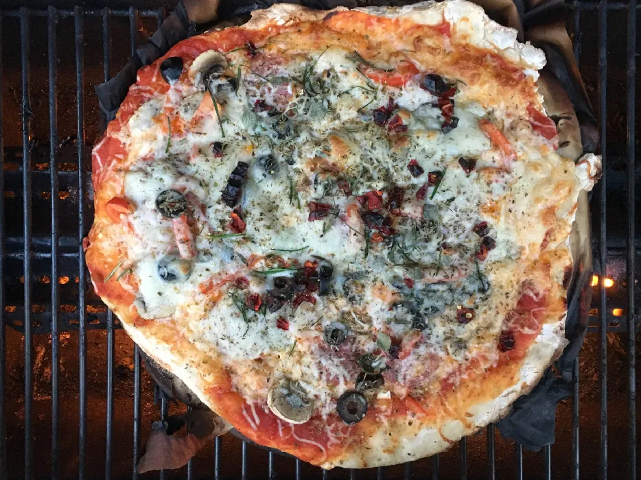 Pizza with tomato sauce, red bell peppers, mushrooms, black olives, salami, and cheese, and spiced with red chili pepper, chives, oregano, thyme, rosemary, and basil, being cooked on a grill.