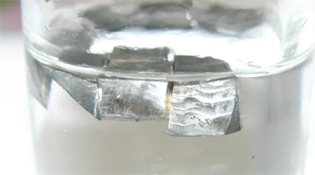 A glass container that is half filled with a colorless liquid is shown. Blocks of a shiny silver solid float on top of the liquid in the container.