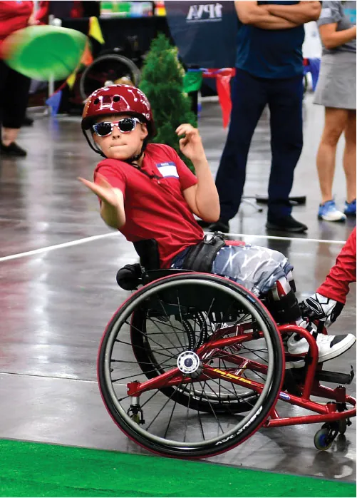 Photo of child in a wheelchair, wearing a helmet and sunglasses, tossing a frisbee.