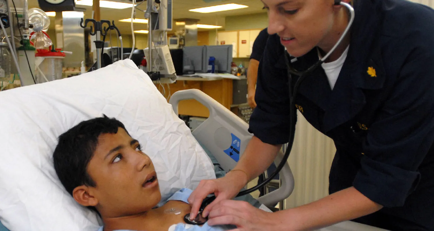 A child client lies on a hospital bed while a nurse checks their heartbeat with a stethoscope.