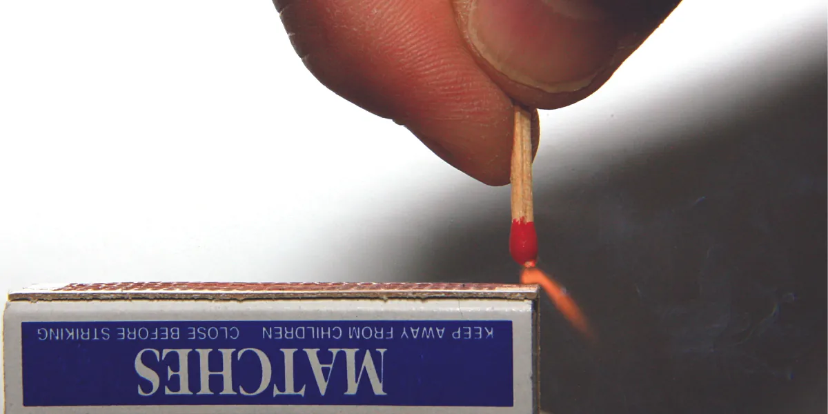 A match held in a person’s hand is ignited as it is scratched along the rough surface of a match box.