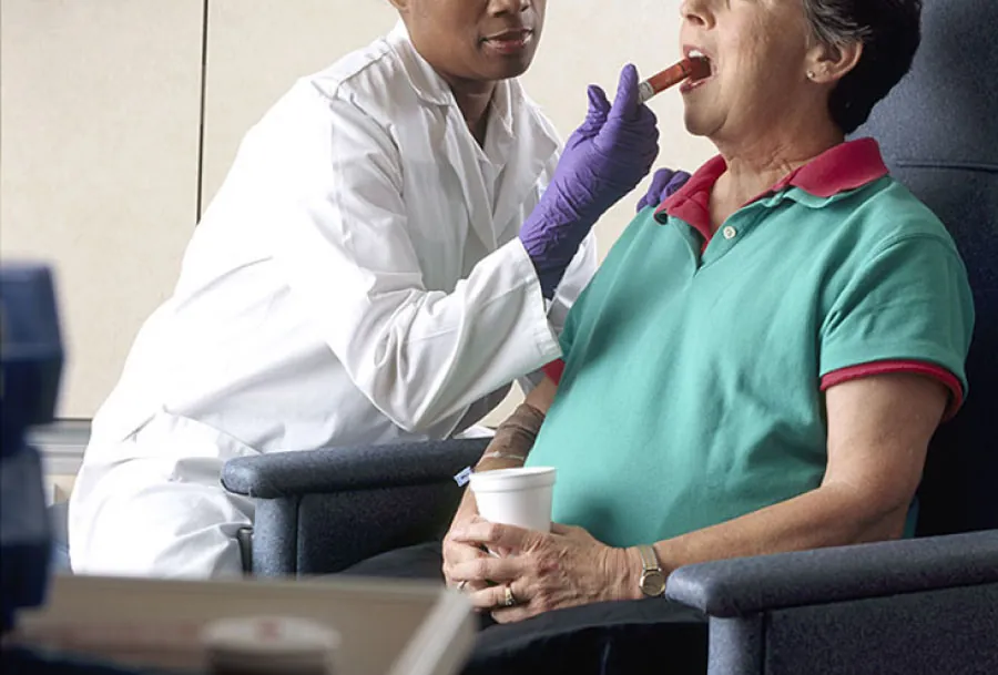 Photo of medical personnel sitting next to seated patient and administering liquid medication through a syringe into the side of the patient’s mouth.