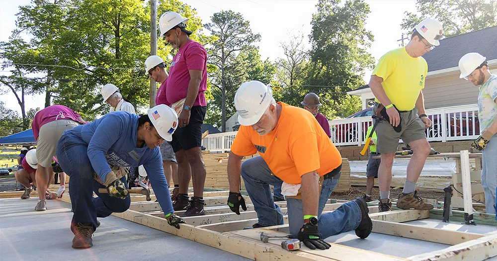 Several people in hard hats and wearing work gloves, with tools nearby and hanging from belts. They are assembling a frame made of wooden beams and planks.