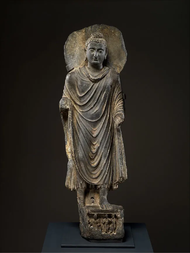 A picture of a golden-black sculpture is shown on a black background. The bare left foot of the man in long flowing robes stands on an ornately carved, square pedestal. His bare right foot is behind him. He is missing his hands and there is a flat circular disc behind his head. He wears a beaded head dressing with a small dome on top of his head, wears earrings, and has a plump, stoic face.