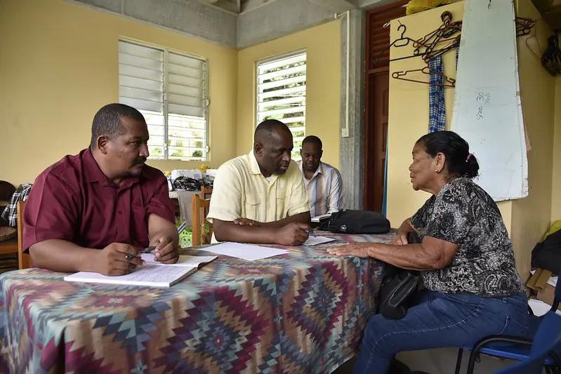 The prime minister of Dominica sits next to an aide at a long table draped in a colorful cloth in a small, informal room, focusing on a constituent sitting across from them.