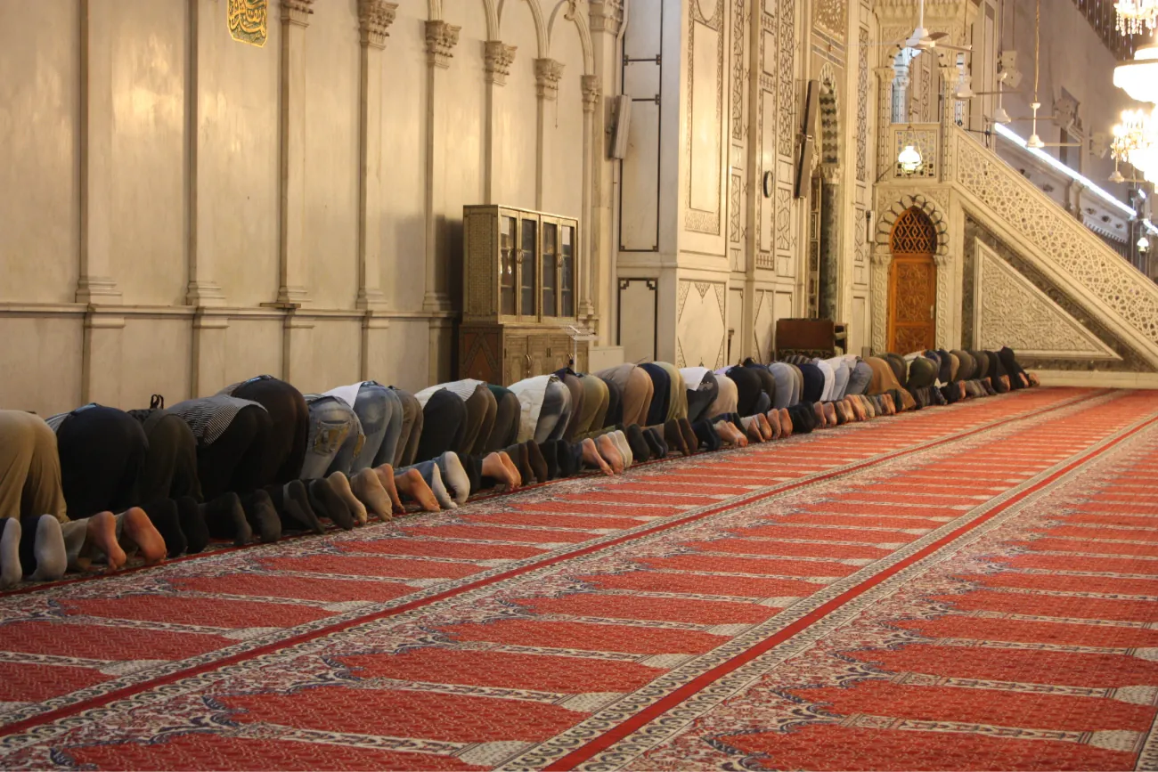 Photo of people kneeling on the floor against a wall of an ornate building, with no shoes on and their heads bowed down to the ground on an ornate carpet.