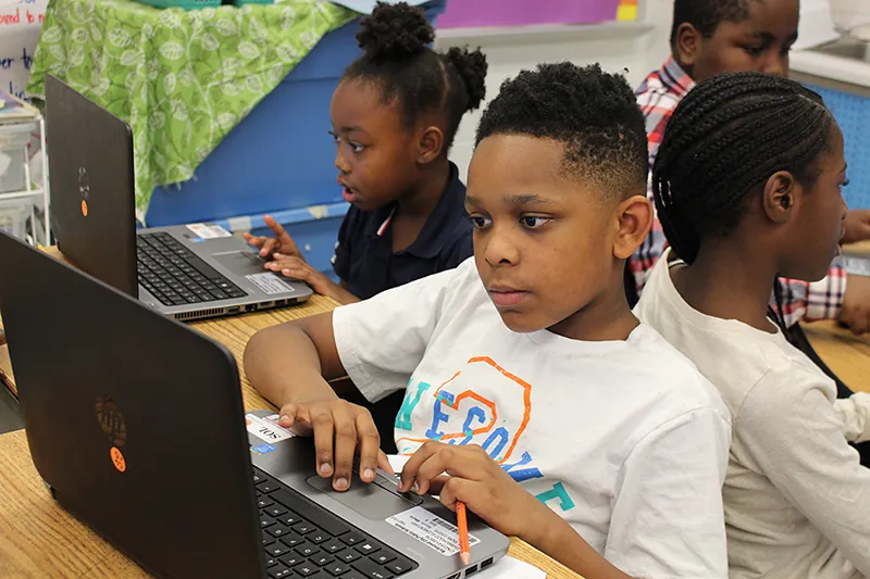 A photo shows several children seated in a classroom working on laptop computers.