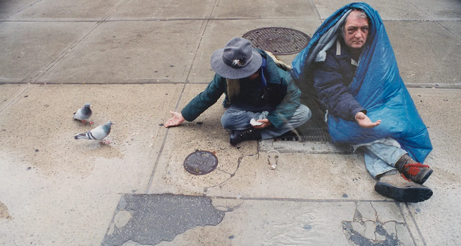 Two people sit on a wet sidewalk. One has a sleeping bag wrapped around their body and over their head, while the other has their hand outstretched towards two pigeons.
