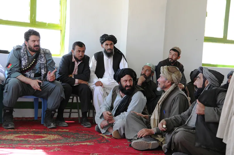A group of people, some wearing turbans, sit together talking  in a room, some on the floor and some in chairs.
