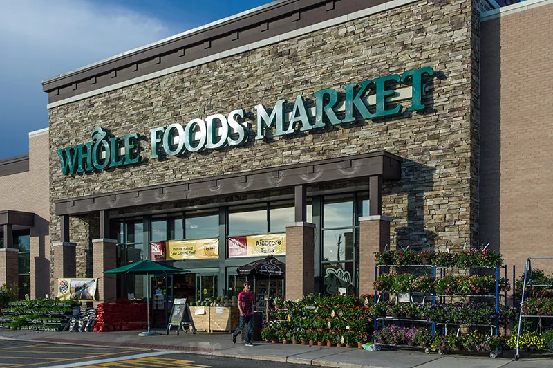 The storefront of a Whole Foods Market has rows of plants to the left and right of the entrance way.