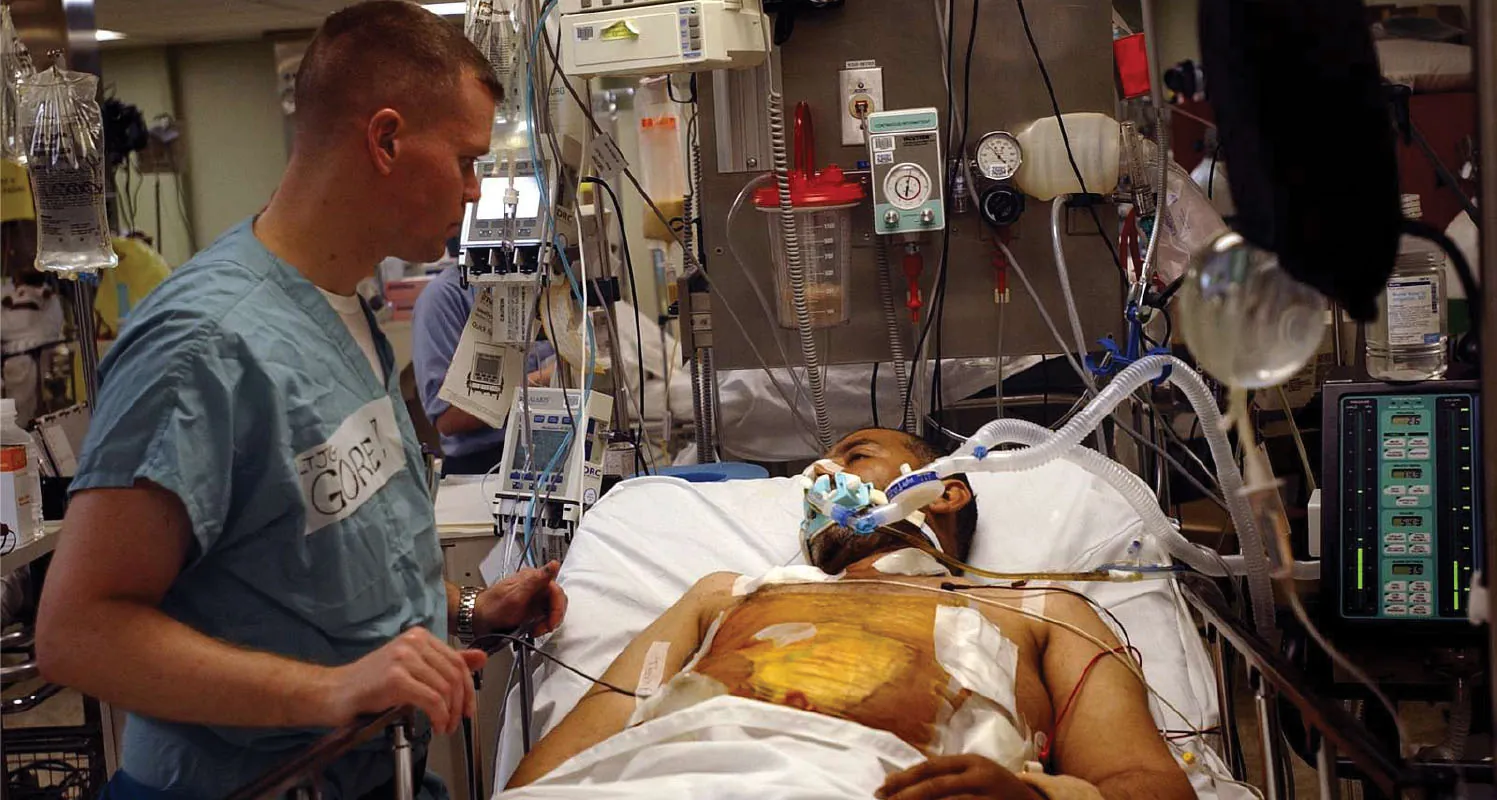 A color photograph of a male nurse and a male patient in the ICU. The patient is shirtless and connected to several monitors and breathing devices. The nurse stands and the bedside looking toward the patient.