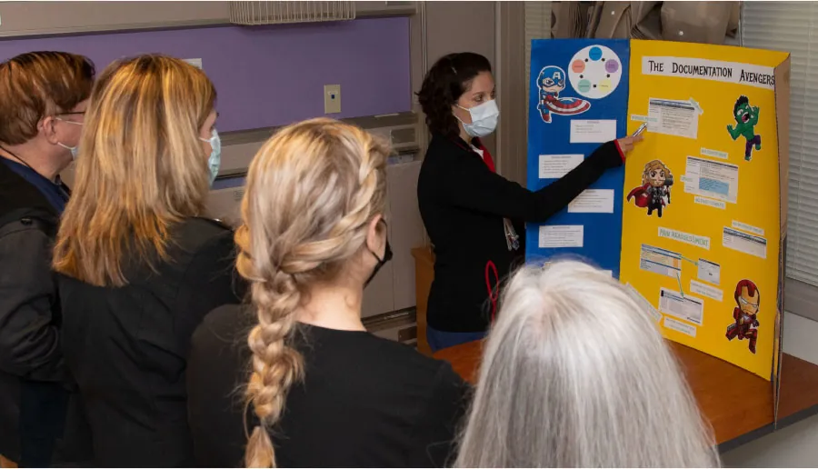 Masked individual stands pointing at tri-fold board while other masked individuals look on.
