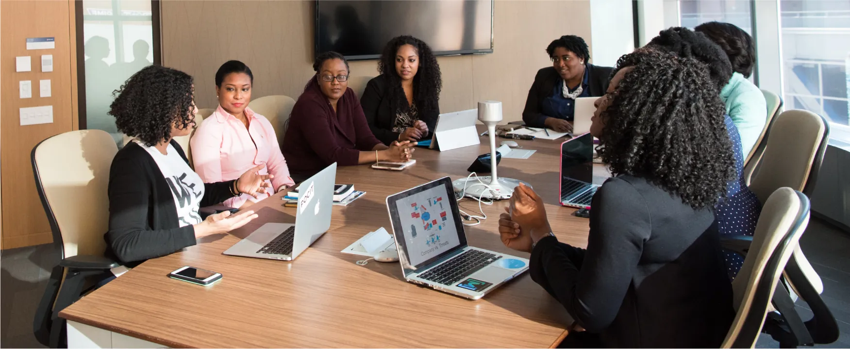 A group of women are shown sitting around a table talking with laptops, tablets, and phones out on the table.
