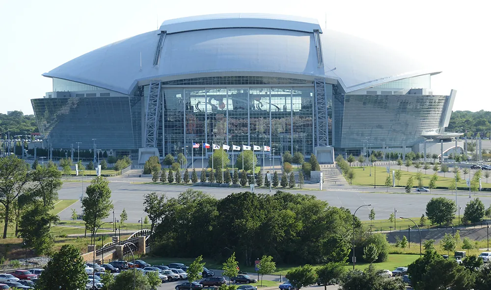 An image of A T and T stadium, a massive steel and glass structure with a domed room, surrounded by parking lots and trees.